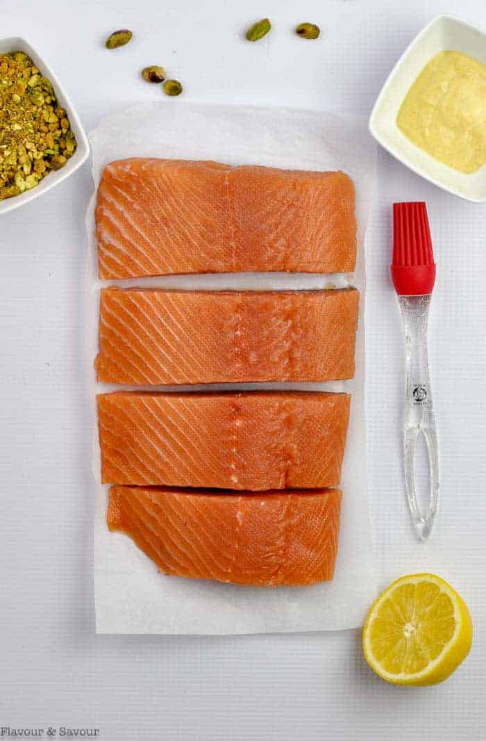 one large salmon fillet cut into 4 pieces with olive oil, a lemon, crushed pistachios and a pastry brush beside.