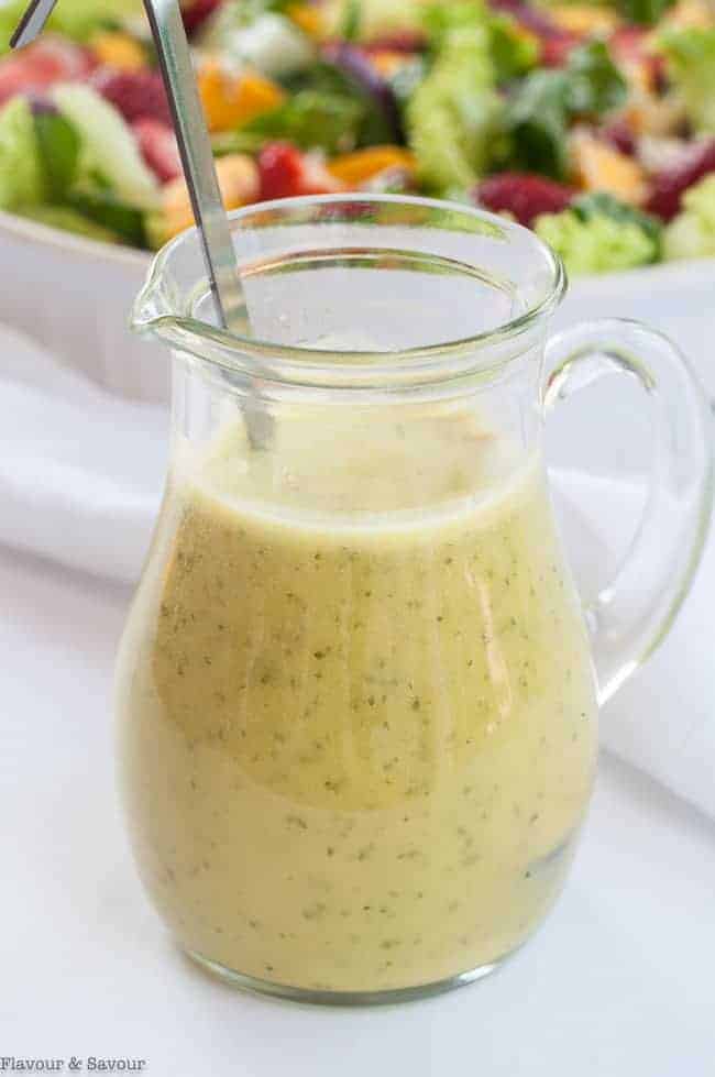 A small pitcher of Everyday Honey Citrus Vinaigrette with salad in the background.