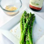 Easy 5 -minute Sesame Asparagus on a plate with a bowl of sesame seeds and a bottle of sesame oil.
