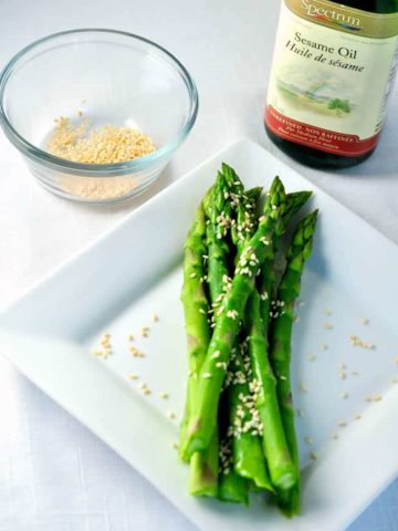 Easy 5 -minute Sesame Asparagus on a plate with a bowl of sesame seeds and a bottle of sesame oil.