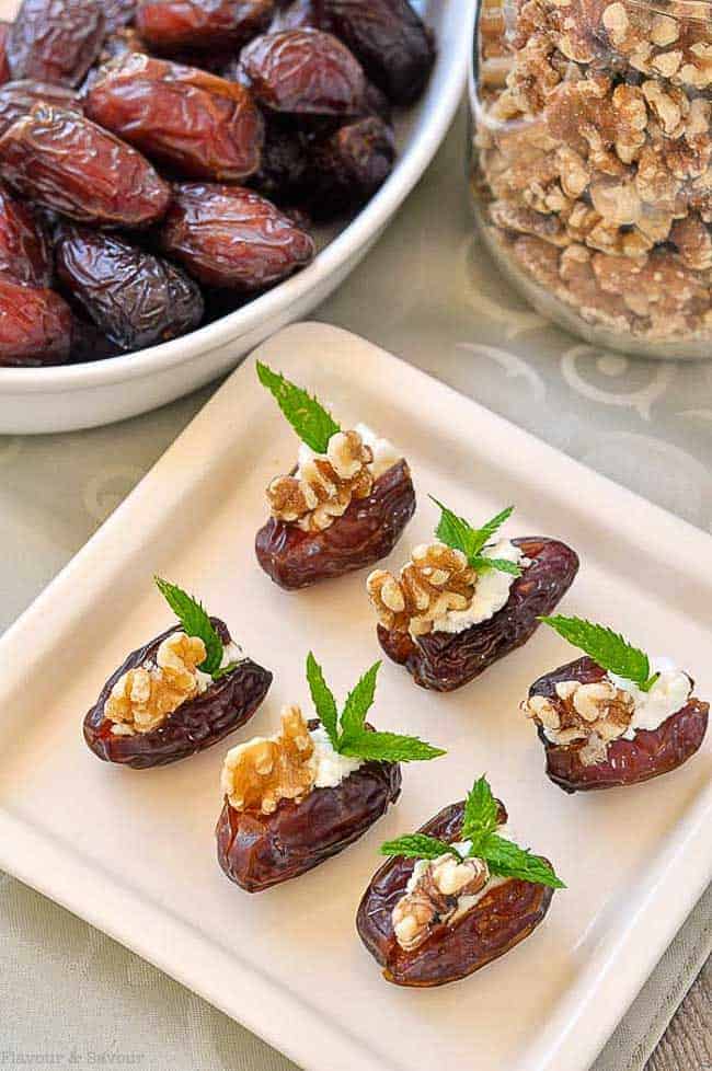 Soft and tender Medjool dates stuffed with creamy goat cheese, toasted walnuts and fresh mint with a jar of walnuts and a bowl of dates in the background.