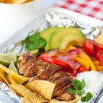 Easy Chicken Fajitas in Foil Packets made with taco-seasoned chicken, peppers, avocado, tomatoes, sour cream and tortilla chips on a checkered cloth.