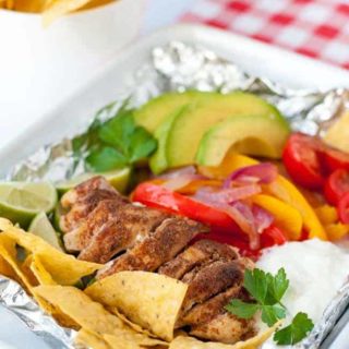 Easy Chicken Fajitas in Foil Packets made with taco-seasoned chicken, peppers, avocado, tomatoes, sour cream and tortilla chips on a checkered cloth.