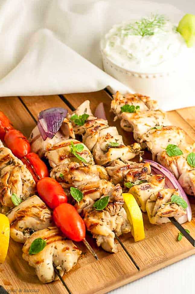Greek Lemon Chicken Kabobs on skewers on a cutting board with skewered tomatoes