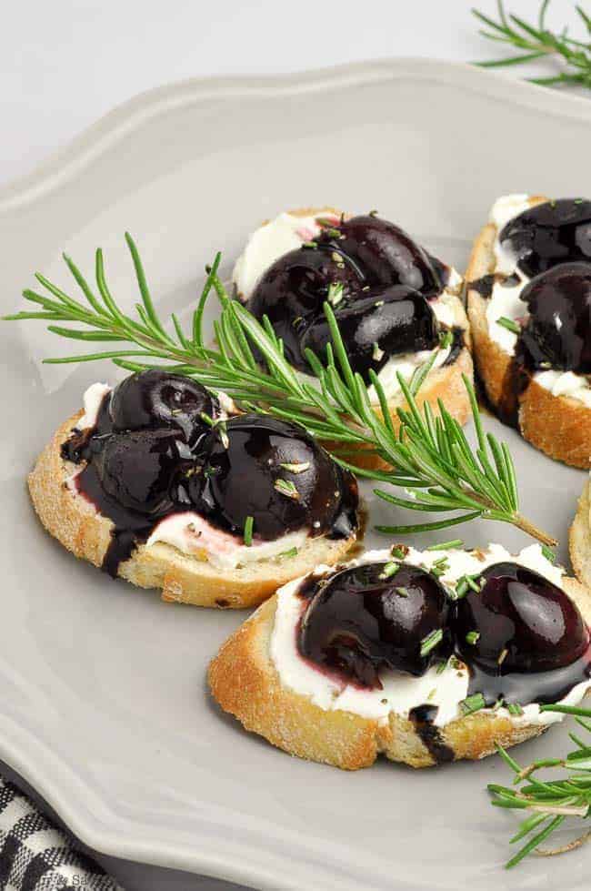 Roasted Cherry Whipped Goat Cheese Crostini appetizers on a plate with a sprig of rosemary
