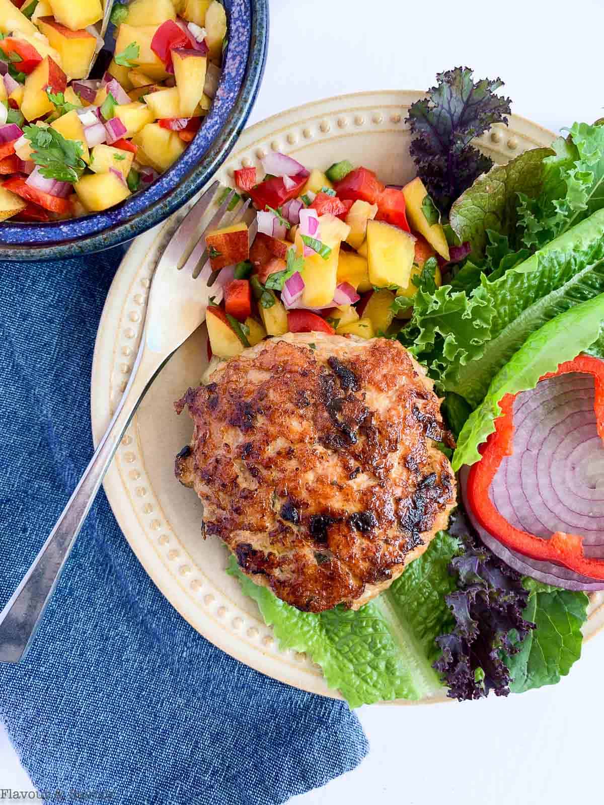 Overhead view of chipotle chicken burger on a plate with peach salsa, greens, red onion and a red pepper ring