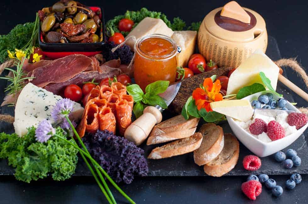 How to Make an Antipasto Platter. An assortment of cheese, charcuterie, olives, fresh vegetables, berries, herbs and edible flowers on a slate tray.