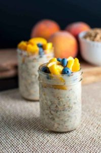 Peach and Blueberry Overnight Oats in 8 oz. Mason jars with fresh peaches in the background.