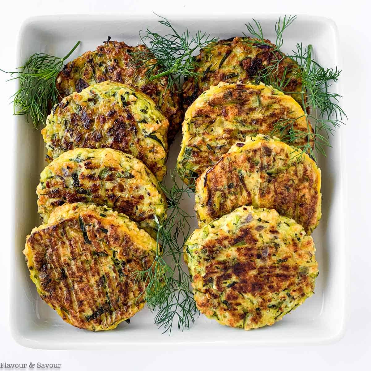 8 gluten-free zucchini patties on a plate with fresh herbs.