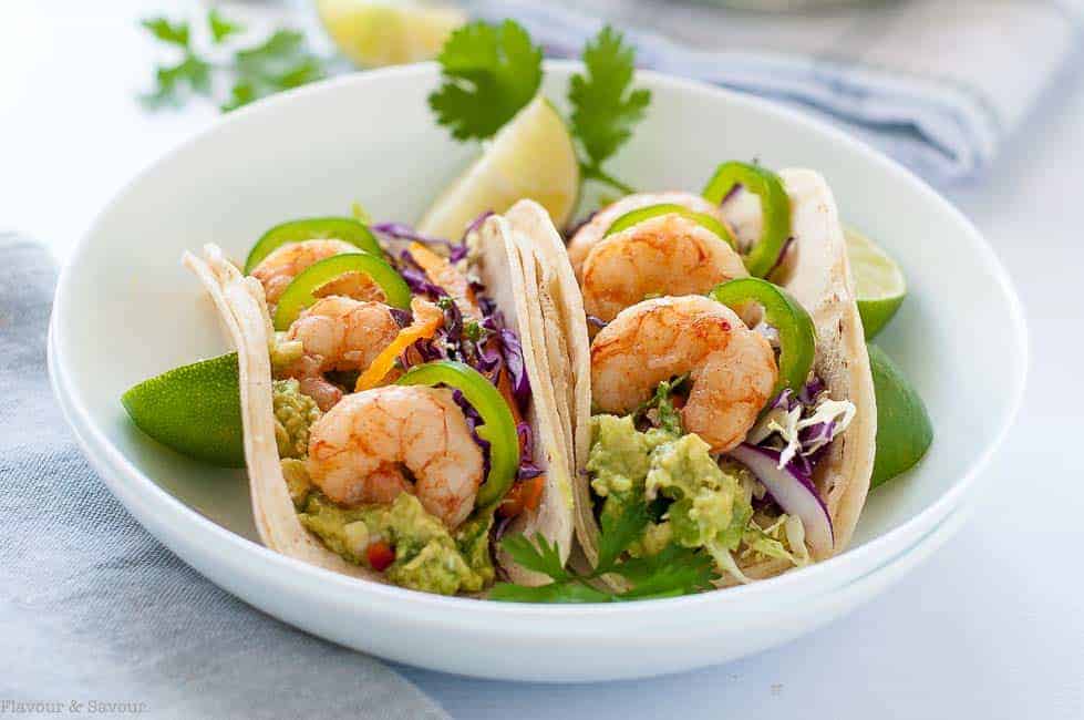 Two Shrimp Tacos with Tomatillo Guacamole and Cilantro Lime Slaw in a shallow white bowl with lime quarters.