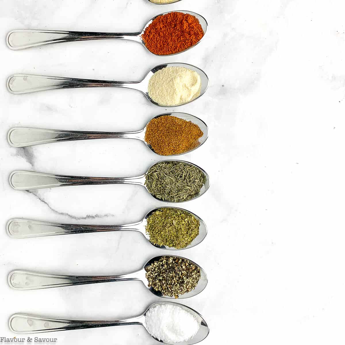 Row of seven silver spoons on white marble, each holding a different spice including paprika, garlic powder and dried herbs