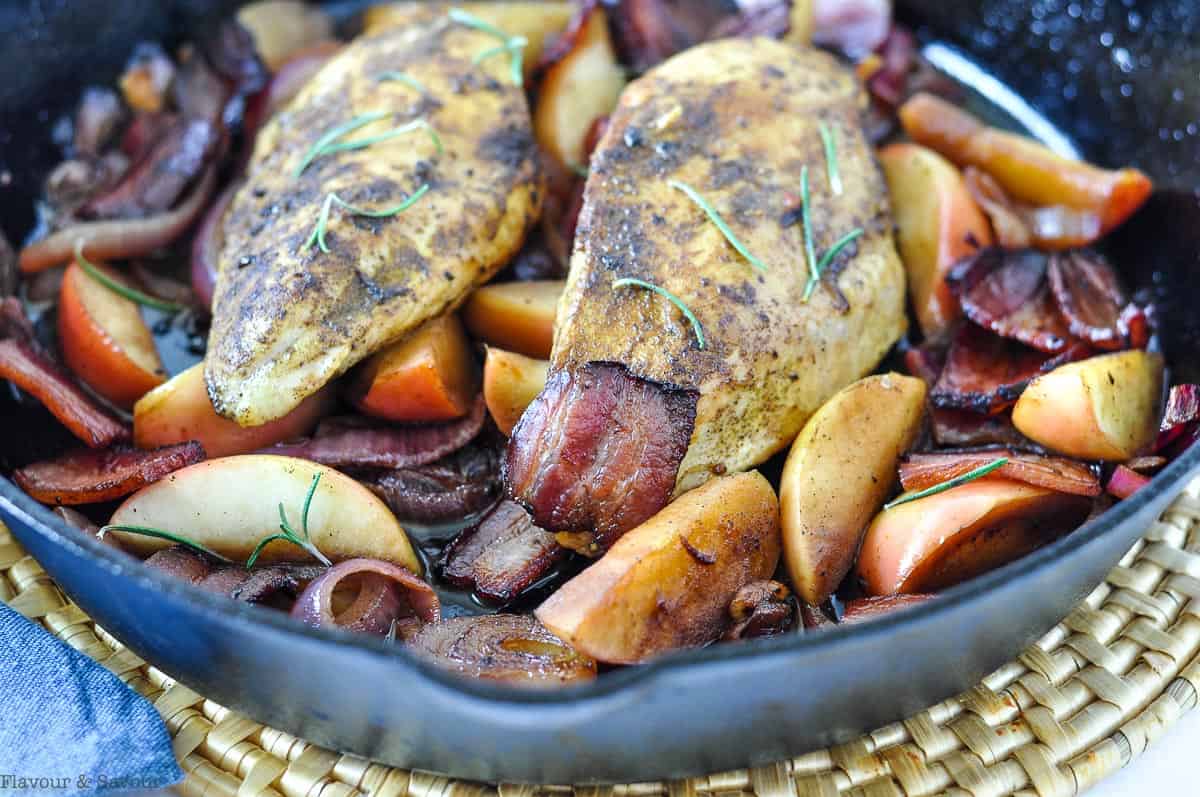 close up view of two cooked chicken breasts in a skillet with apples and bacon