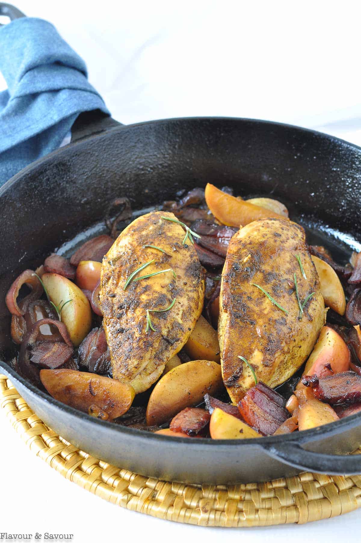 Two cooked chicken breasts in a cast iron skillet with sliced apples and bacon