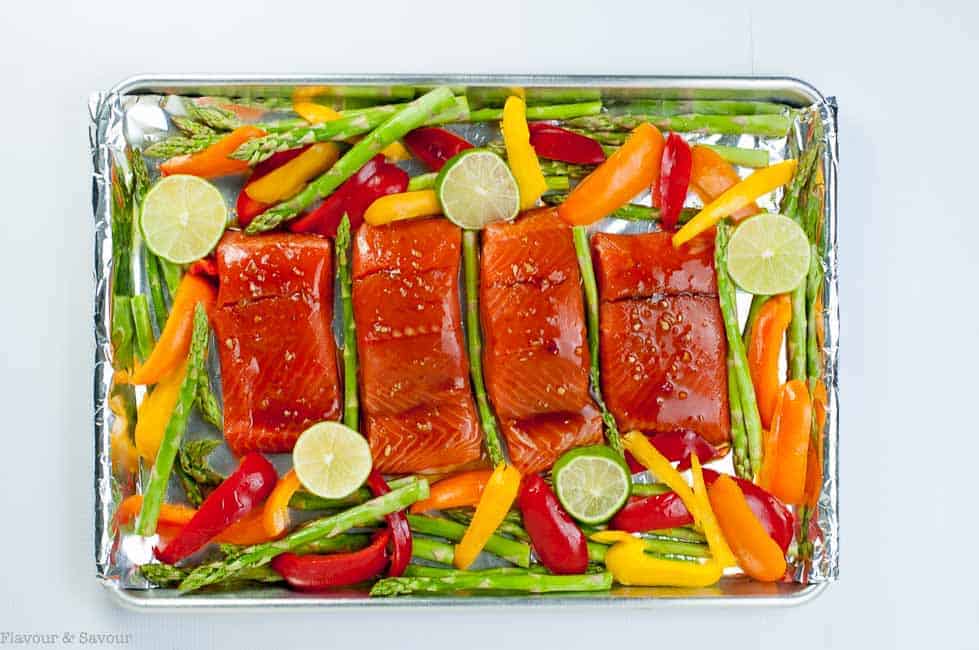 Raw Thai Chili Salmon fillets surrounded by raw asparagus spears and sliced bell peppers on a sheet pan.