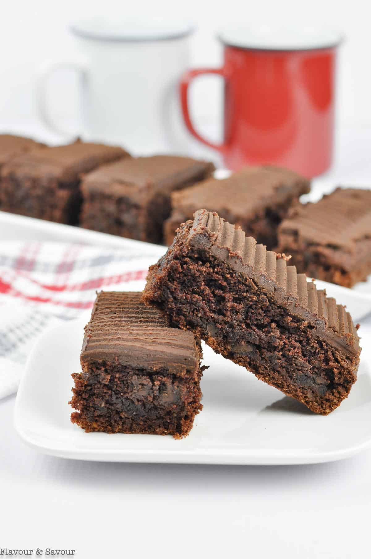 Two bars of Brownies on a plate with coffee cups in the background.