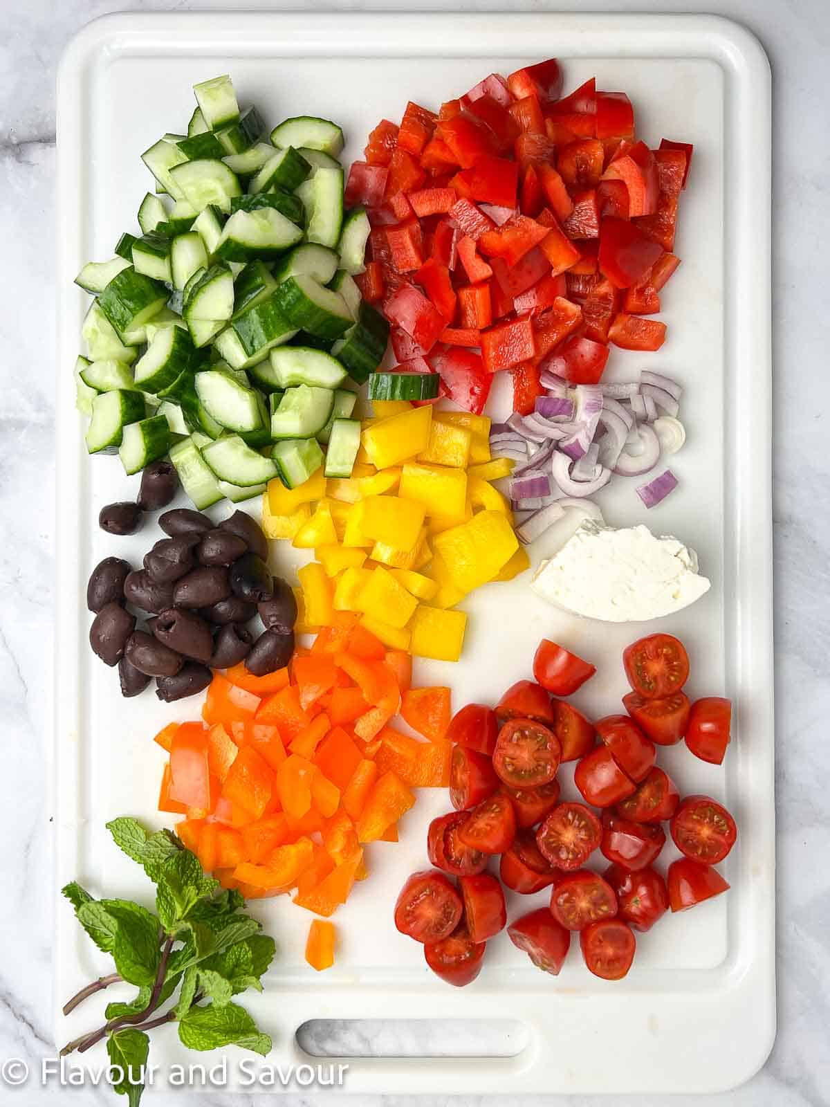 Chopped vegetables including cucumber, bell peppers, red onions, tomatoes as well as olives, feta cheese and mint leaves as ingredients for Greek Chicken Salad.