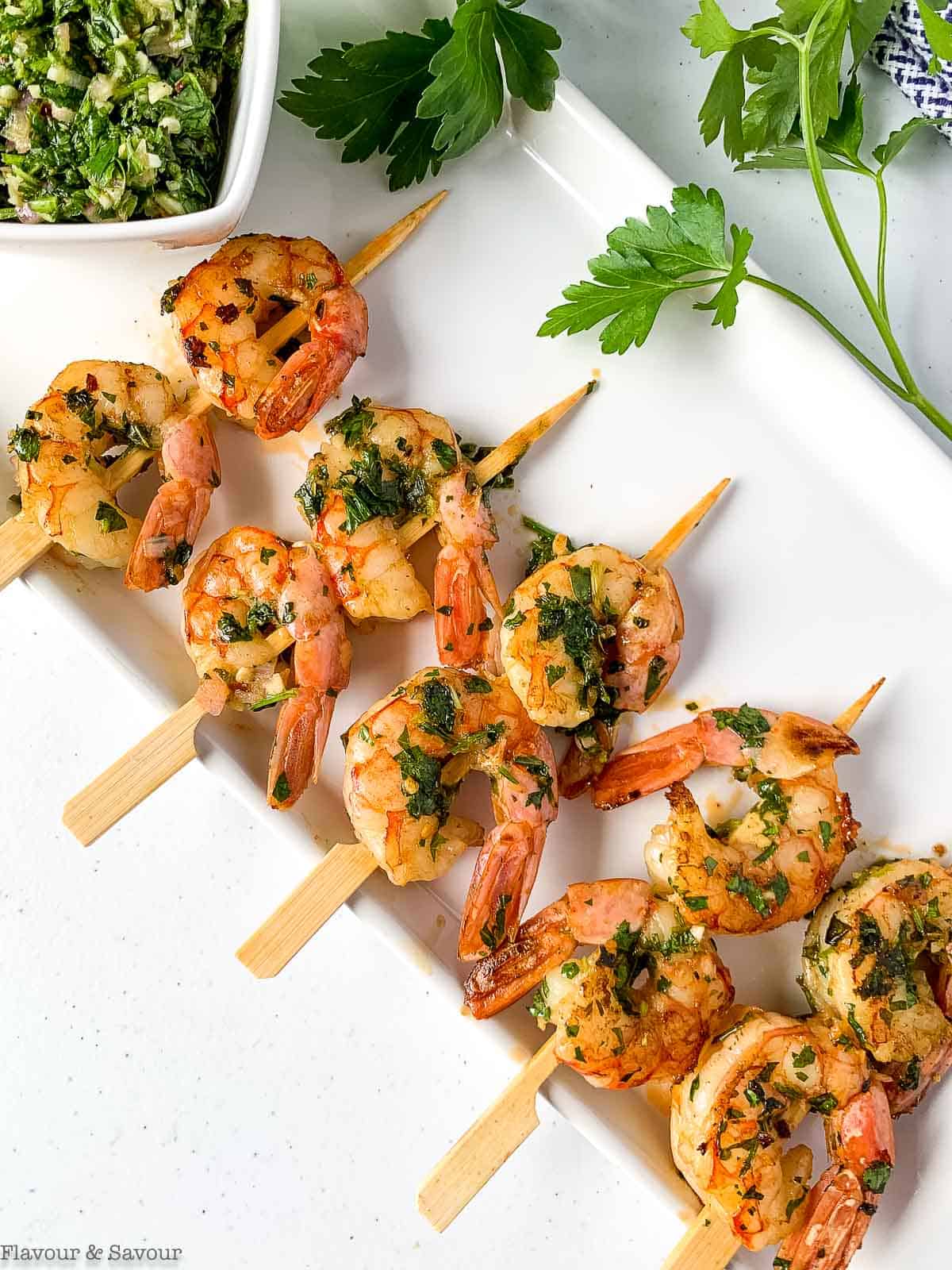 Shrimp skewers with Chimichurri sauce on a white platter.
