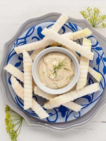 raw kohlrabil sticks on a blue patterned plate with a bowl of Dijon Dilly Dip