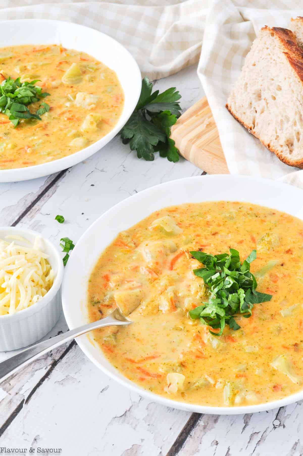 Bowls of Broccoli Cheddar Soup garnished with fresh parsley with a bowl of shredded cheese nearby.