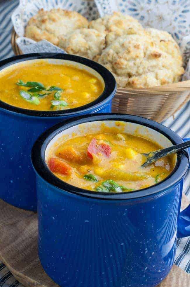 A spoonful of Sweet Potato Corn Chowder from a blue soup mug with a basket of baking powder biscuits in the background.