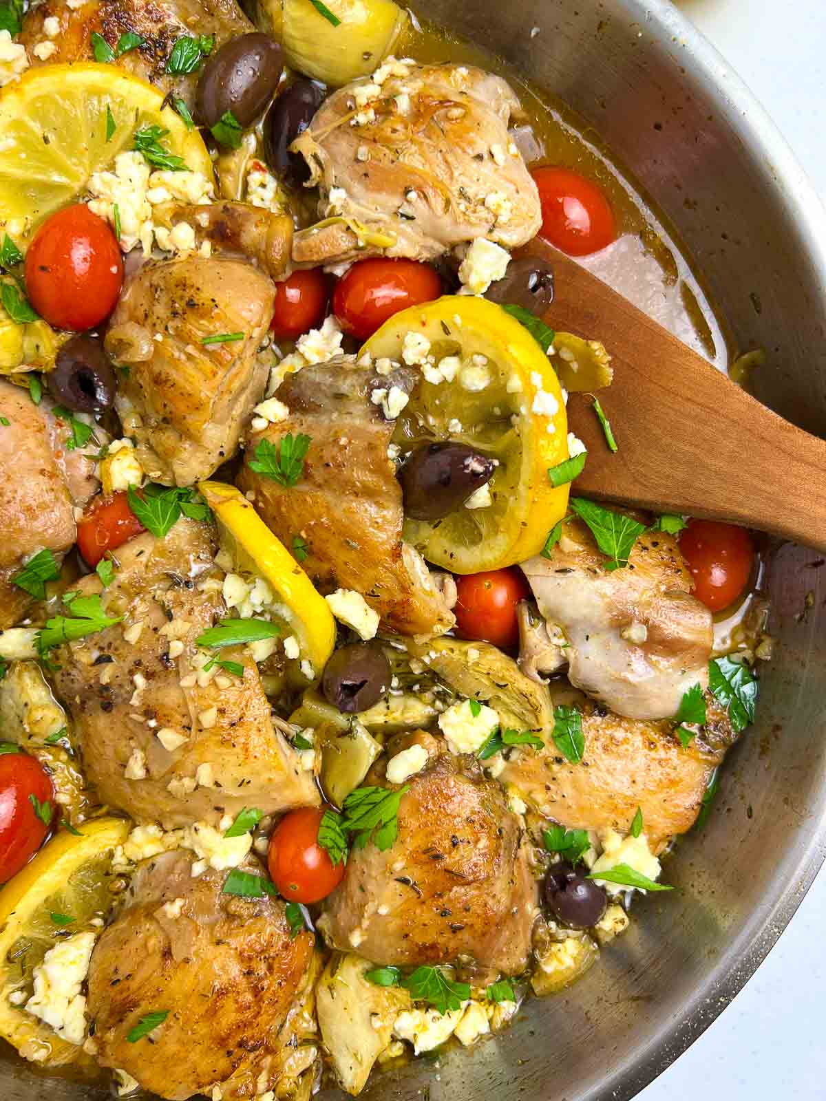 Close up view of baked lemon artichoke chicken in a skillet with a wooden spoon.