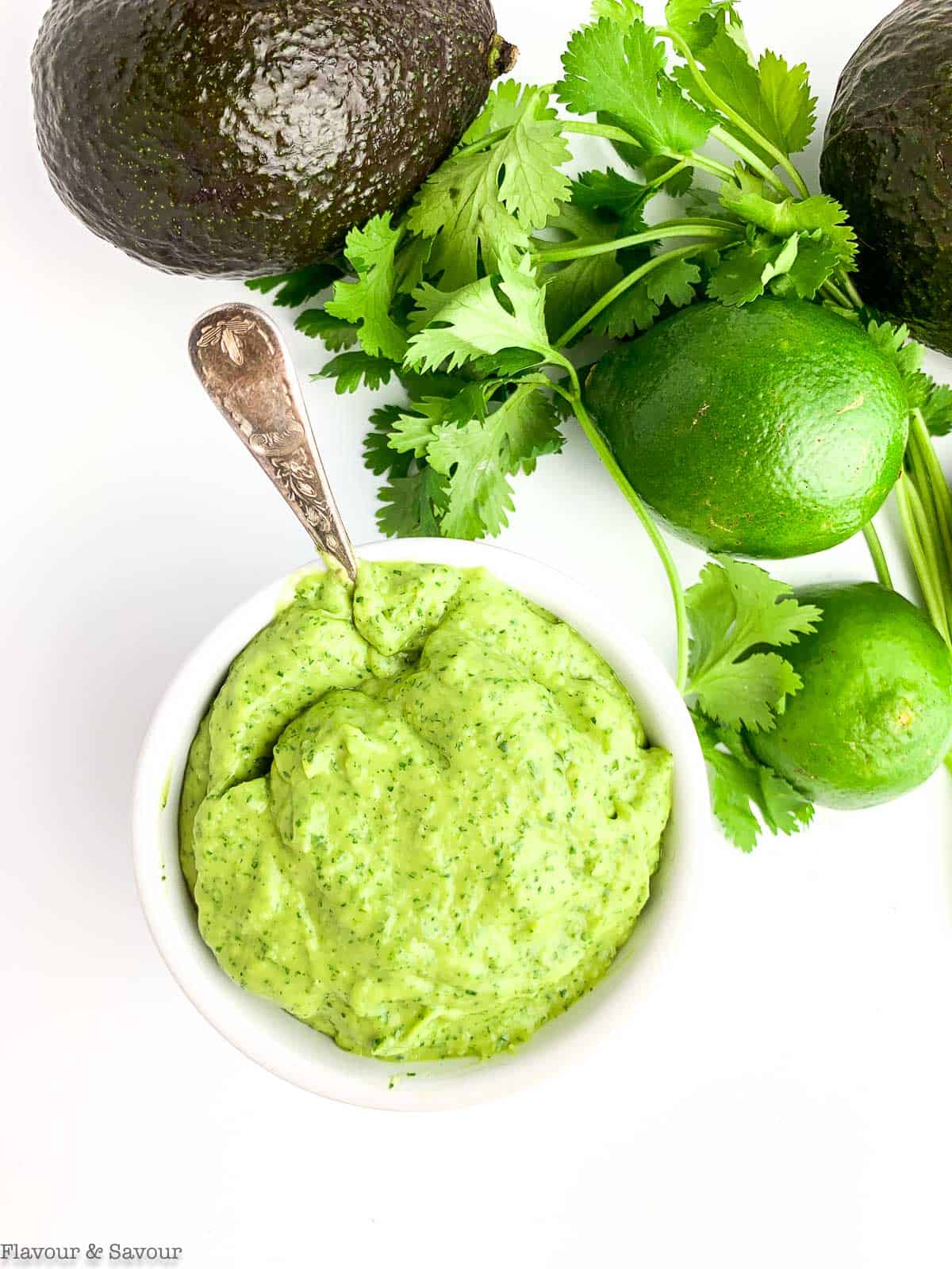 Overhead view of a small bowl of avocado cream with avocados, lime and cilantro beside it.
