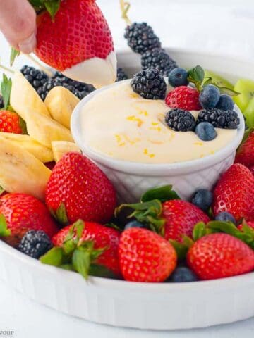 A hand dipping a strawberry into a small bowl of lemon curd fruit dip.