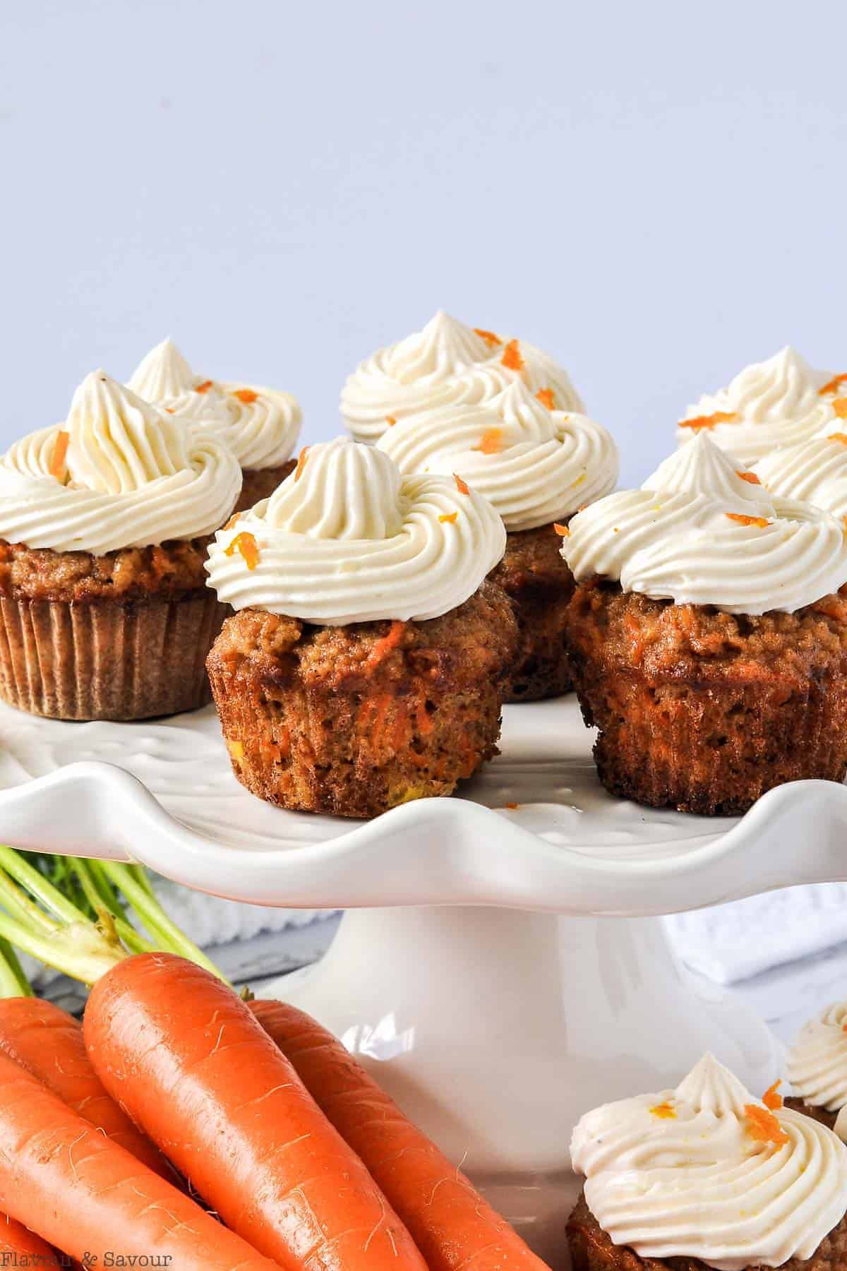 Mini Gluten-Free Carrot Cake Cupcakes on a white pedestal with fresh carrots in the foreground.