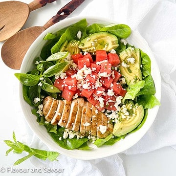 A bowl of greens with sliced chicken breast, watermelon cubes, avocado slices and feta cheese.