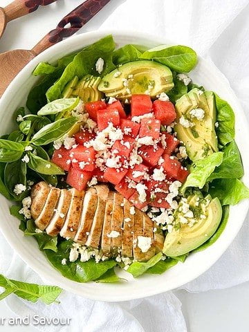 A bowl of greens with sliced chicken breast, watermelon cubes, avocado slices and feta cheese.