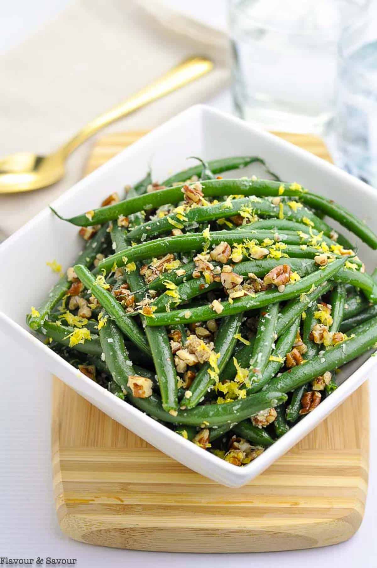 A square bowl of green beans garnished with pecans and Parmesan cheese.