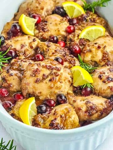 A serving bowl with chicken thighs baked with cranberries and rosemary.