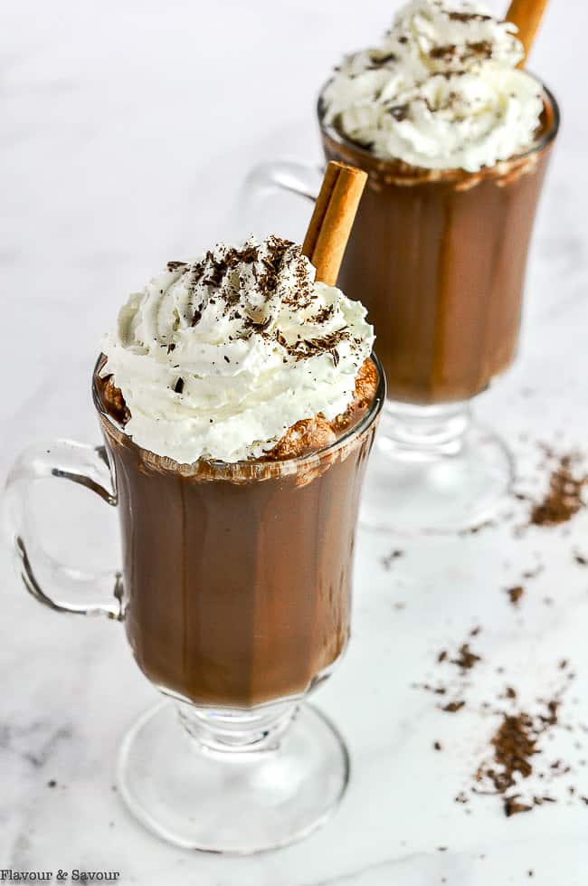 Two glass mugs of hot chocolate with whipped cream, chocolate shavings and cinnamon sticks.