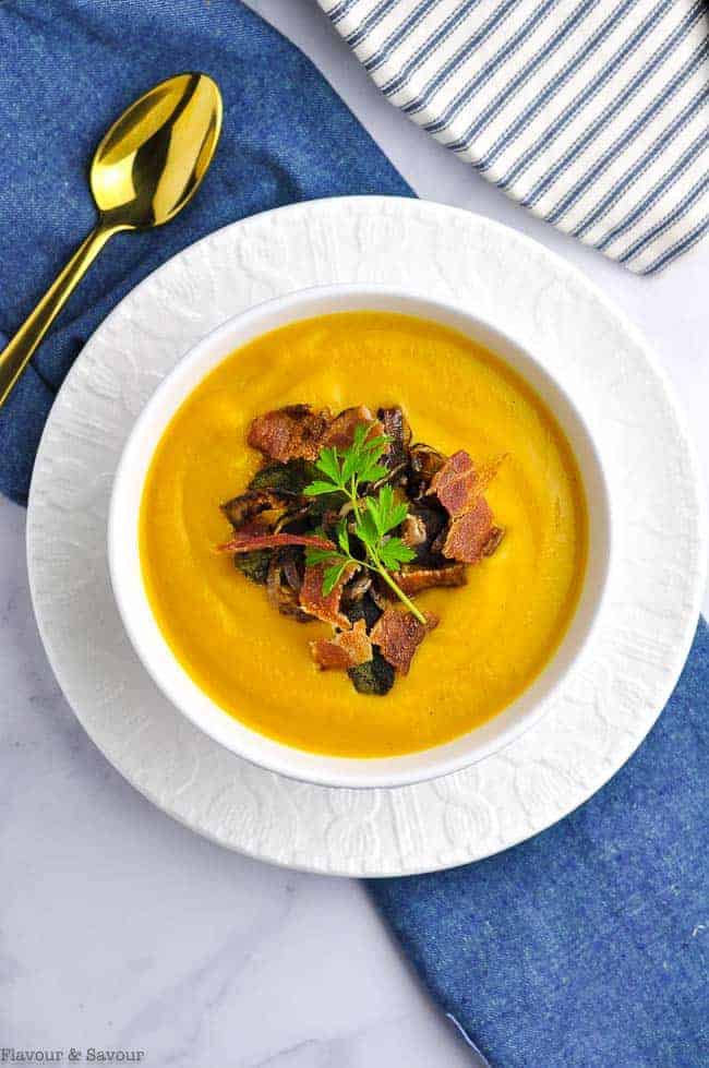 Butternut Squash Soup topped with crispy fried shallots, toasted sage leaves and bacon bits in a white bowl on a blue cloth.
