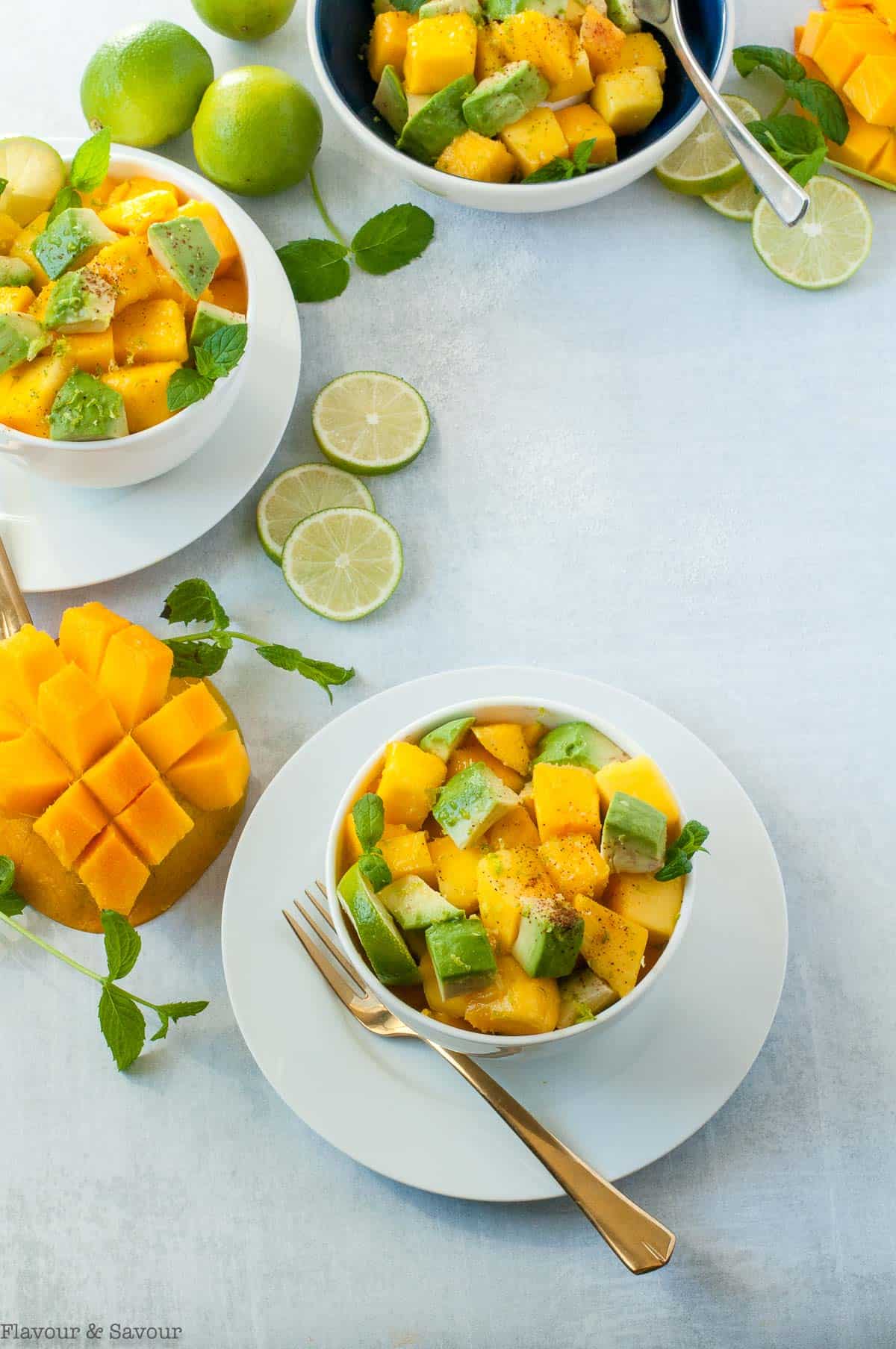 three bowls of Mango Avocado Chili Lime Salad with lime slices, mint leaves and cubed mango on the table