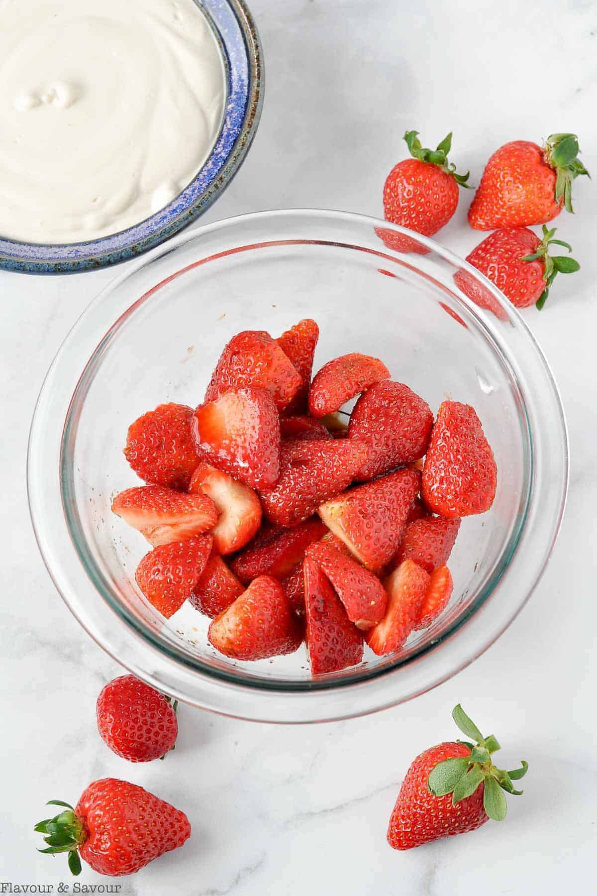 A bowl of halved strawberries and a bowl of cashew cream with a few fresh strawberries on the counter top.