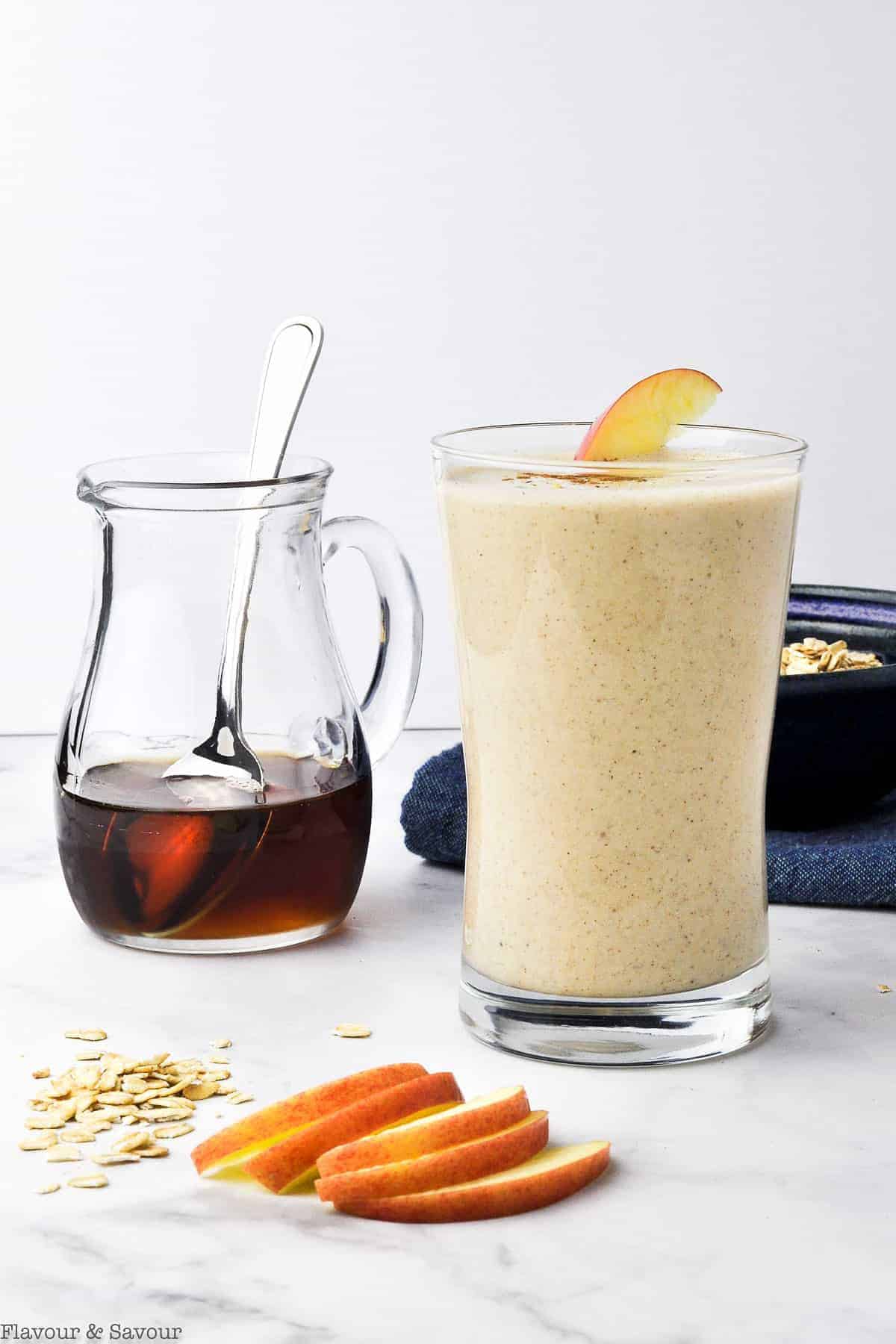 Apple oatmeal breakfast smoothie with apple slices and a pitcher of maple syrup nearby.