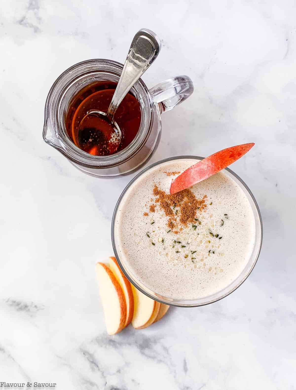 Overhead view of a glass with apple cinnamon smoothie and a small pitcher of maple syrup.