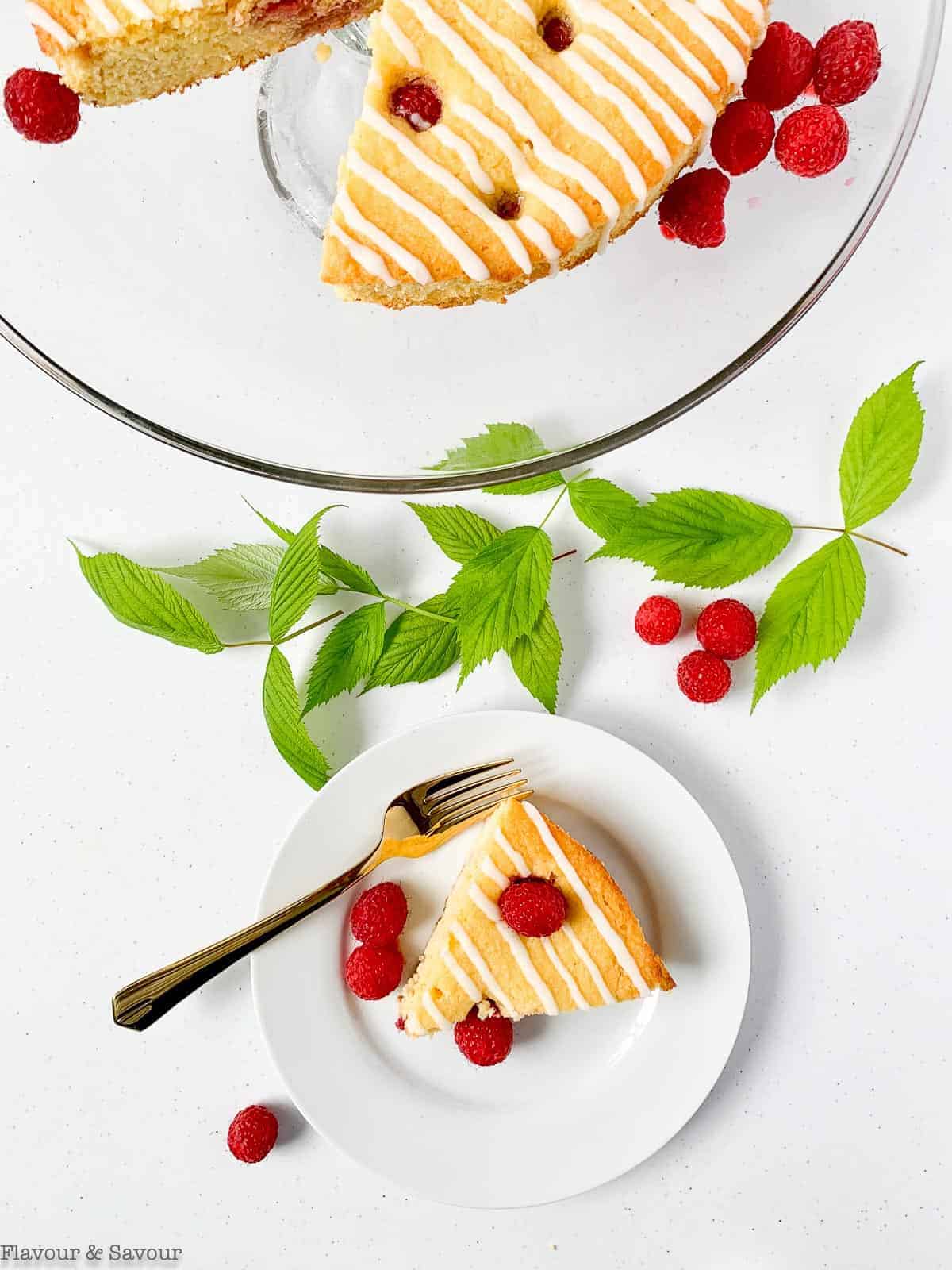 A slice of Raspberry Lemon Ricotta Cake with the rest of the cake on a clear glass pedestal