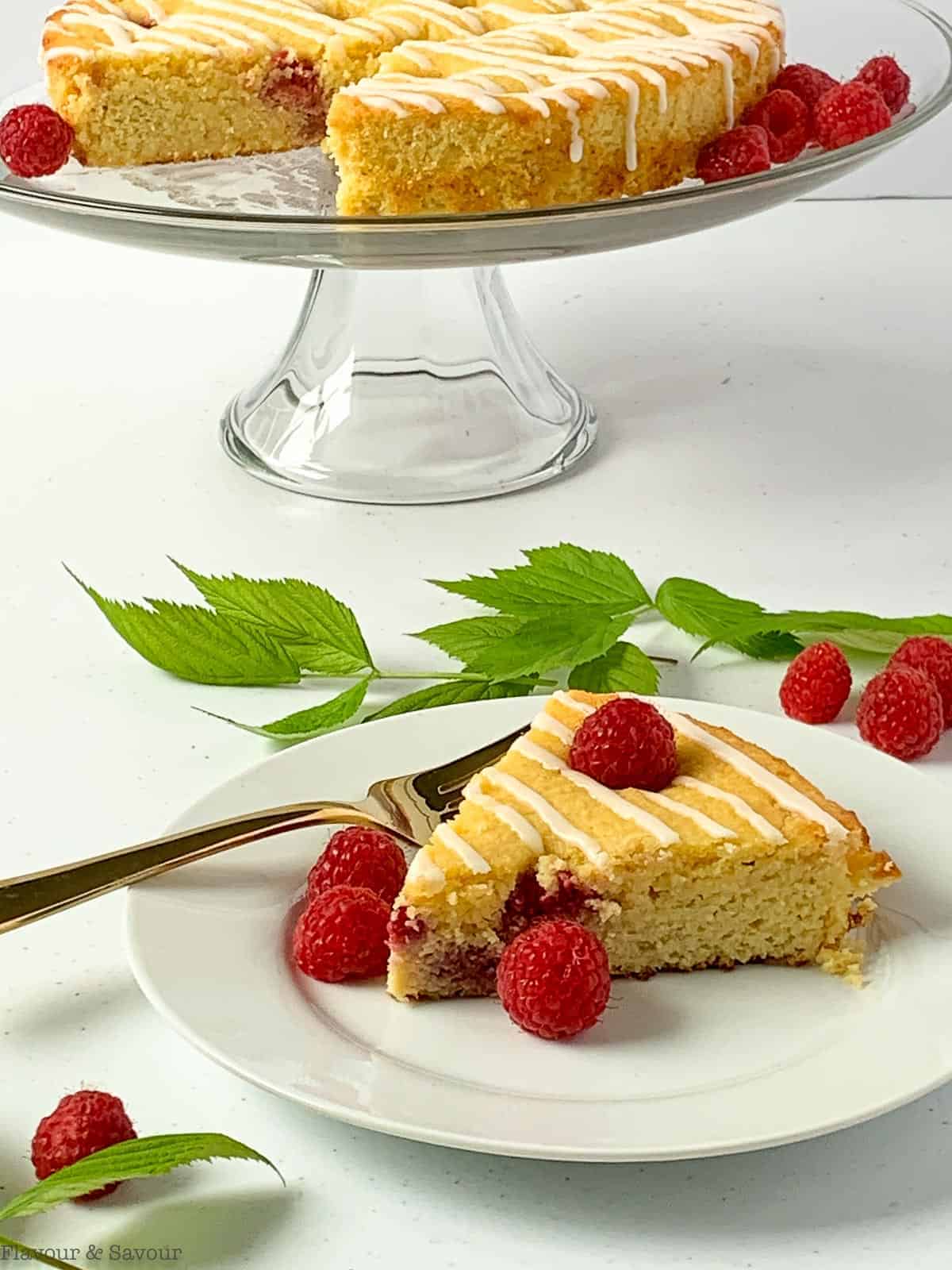 A slice of Raspberry Lemon Ricotta Cake on a white plate with the cake on a glass pedestal stand behind.