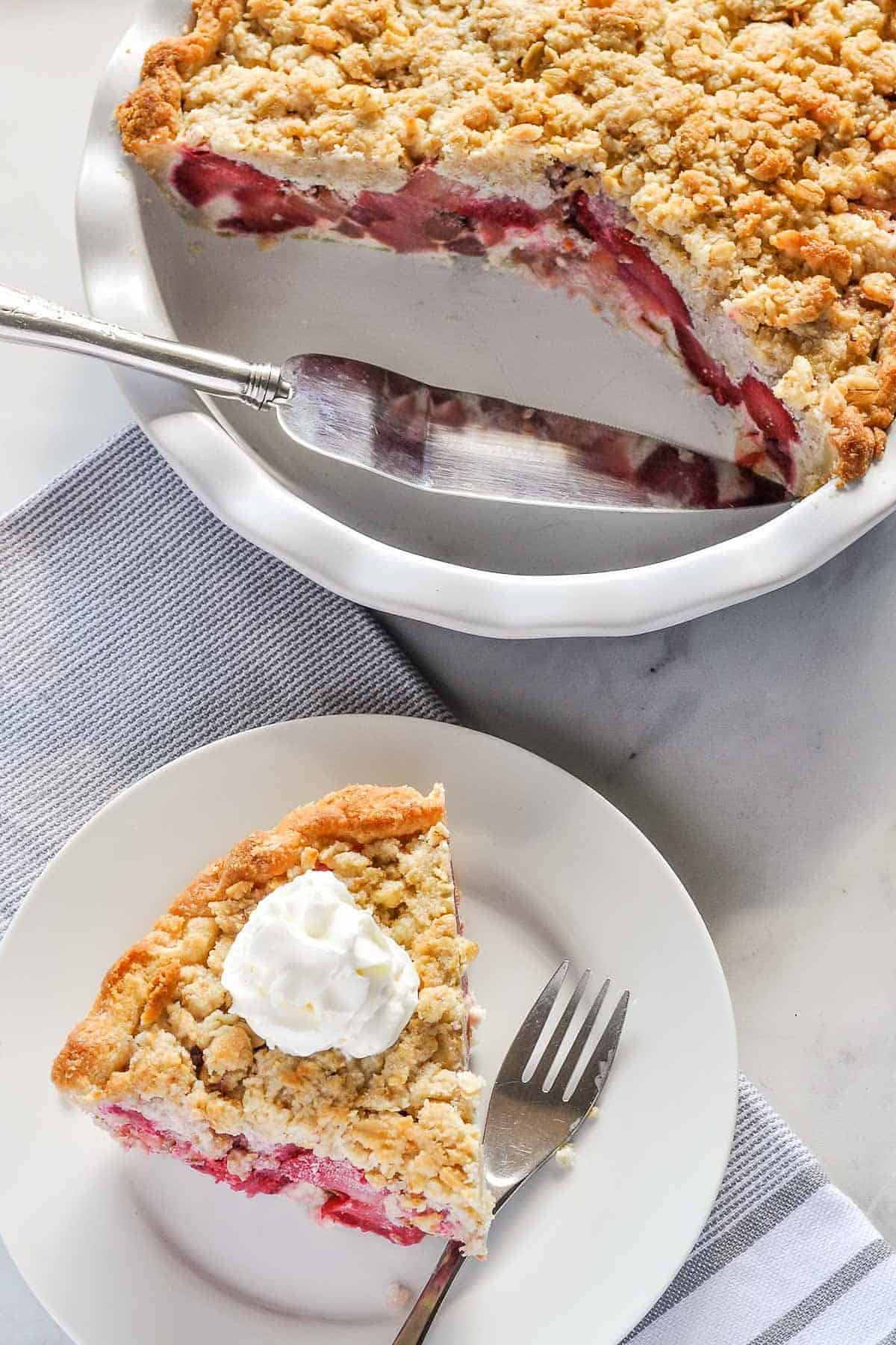 A slice of Strawberry Rhubarb Crumble Pie with a pie plate and the remainder of the pie beside it.