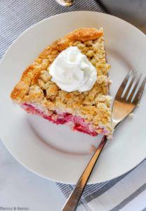 A slice of Strawberry Rhubarb Crumble Pie topped with whipped cream on a white plate with a fork.