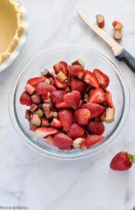 Sliced strawberries and chopped rhubarb in a bowl beside an empty unbaked pie crust.