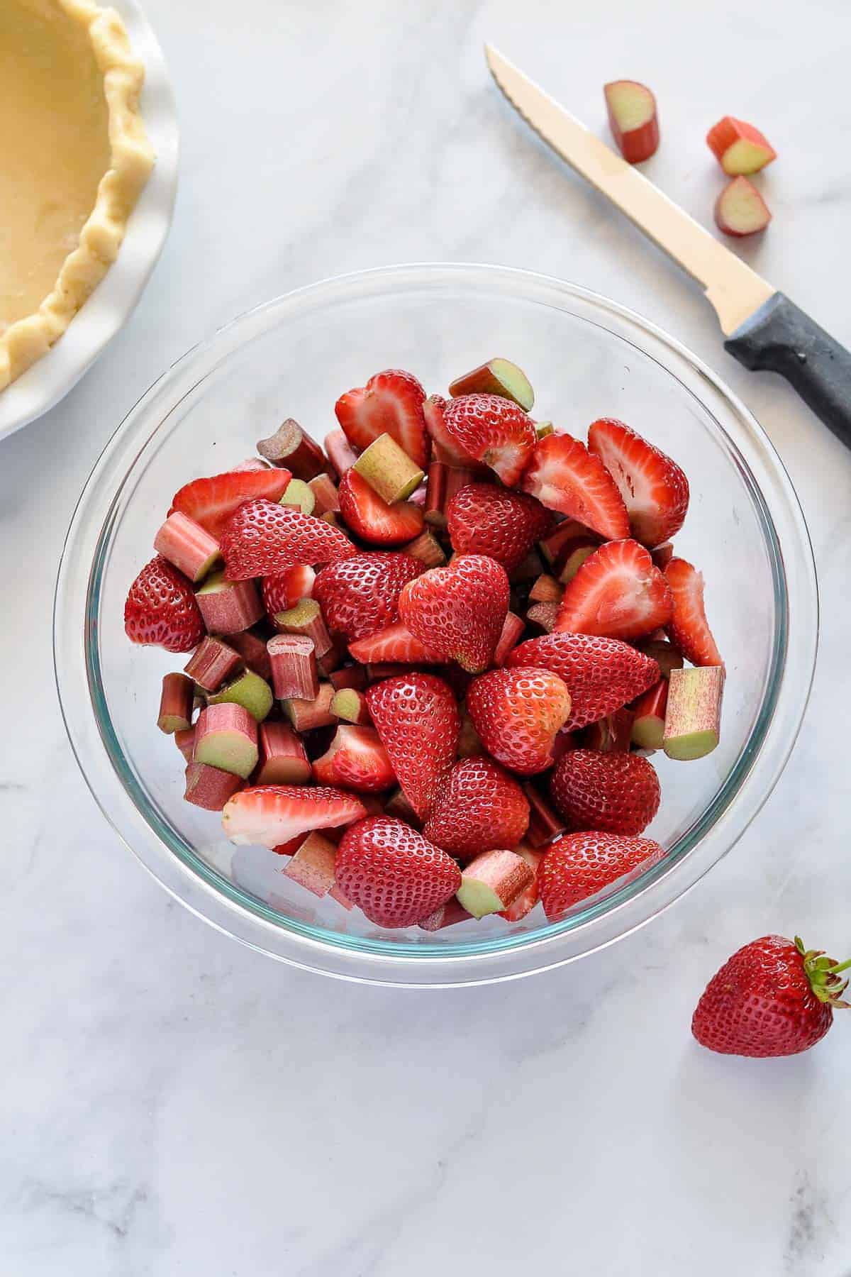 Sliced strawberries and chopped rhubarb in a bowl beside an empty unbaked pie crust.