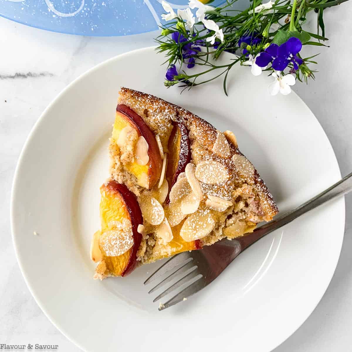 A slice of Gluten-Free Flourless Nectarine Ricotta Cake on a white plate with fresh blue and white flowers