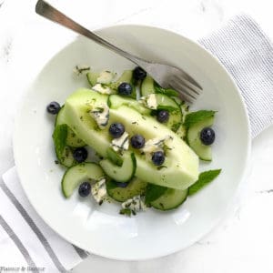a shallow bowl with a wedge of honeydew melon with cucumbers, blueberries, blue cheese and mint leaves