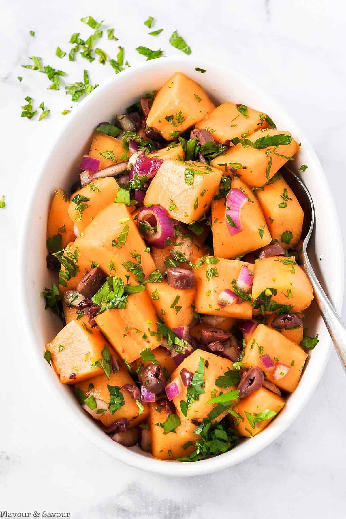 Overhead view of an oval bowl of Mediterranean Cantaloupe Salad with Olives and Mint.