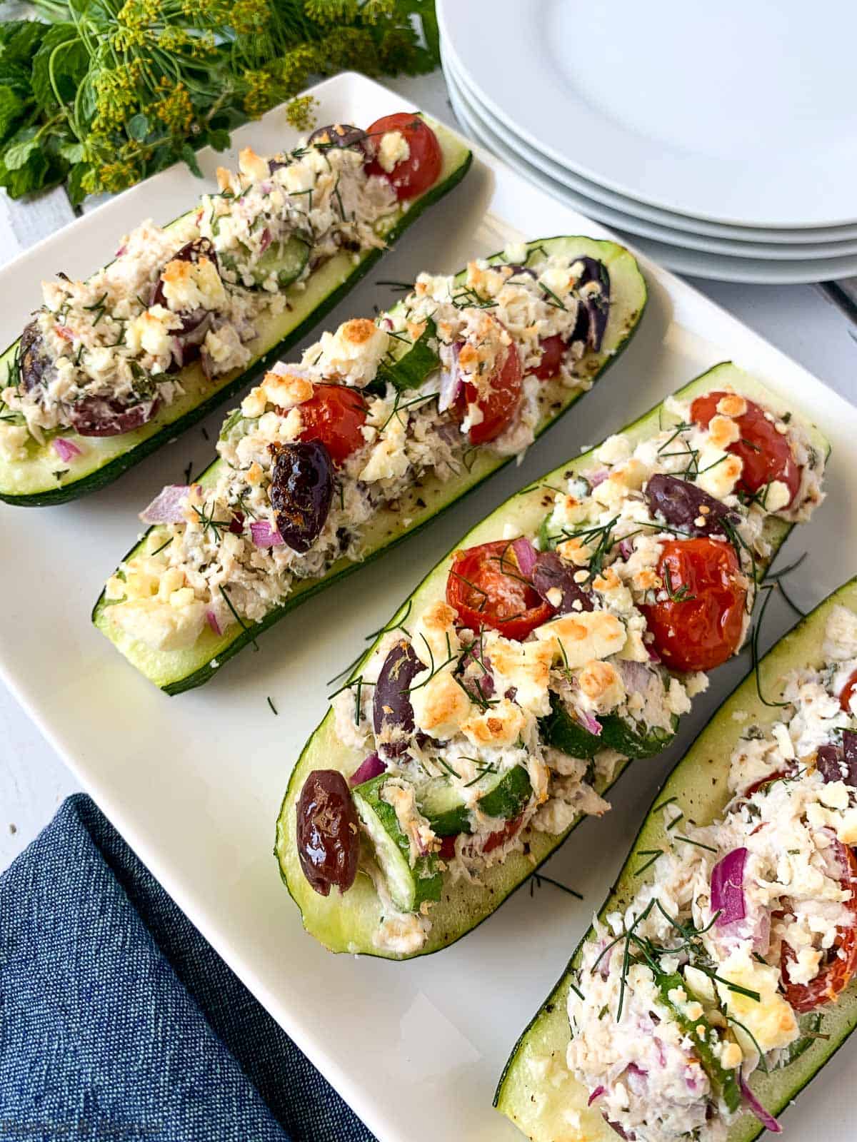 Greek Chicken Stuffed Zucchini Boats on a white platter with fresh herbs in the background
