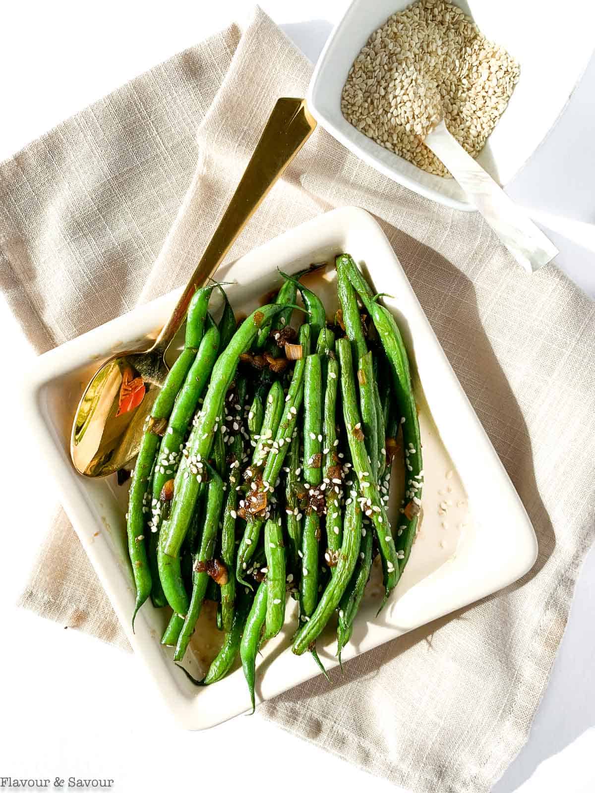 Sesame ginger green beans in a square dish with a bowl of sesame seeds.