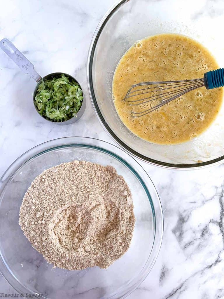 Two bowls, one with wet ingredients and one with dry ingredients and a cup measure of grated zucchini